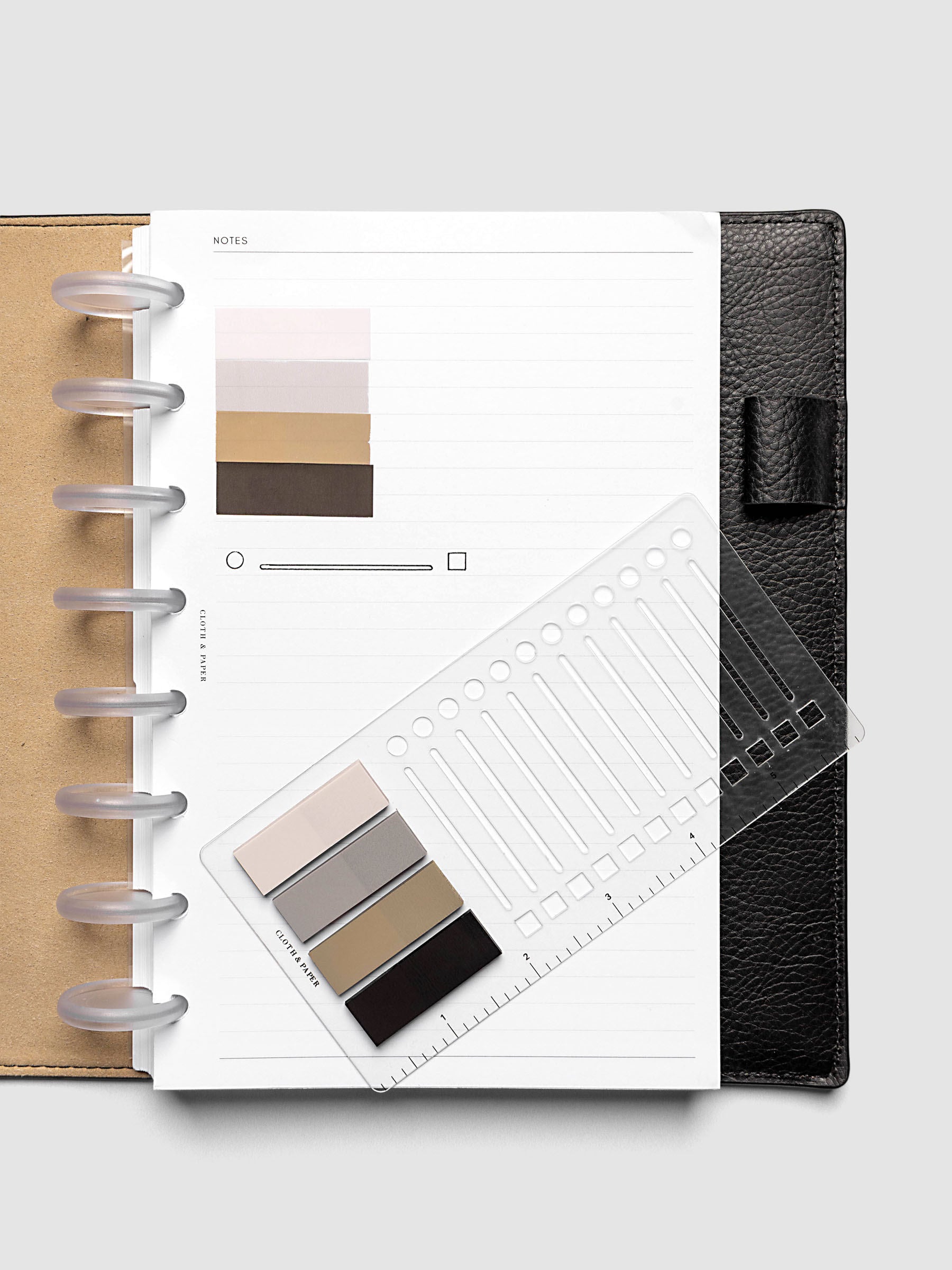 Stencil Page Flag Set shown in use inside an open discbound planner, with stencil laid across a lined notes page and page flags fanned on the page above it; black pebbled leather planner cover visible at right