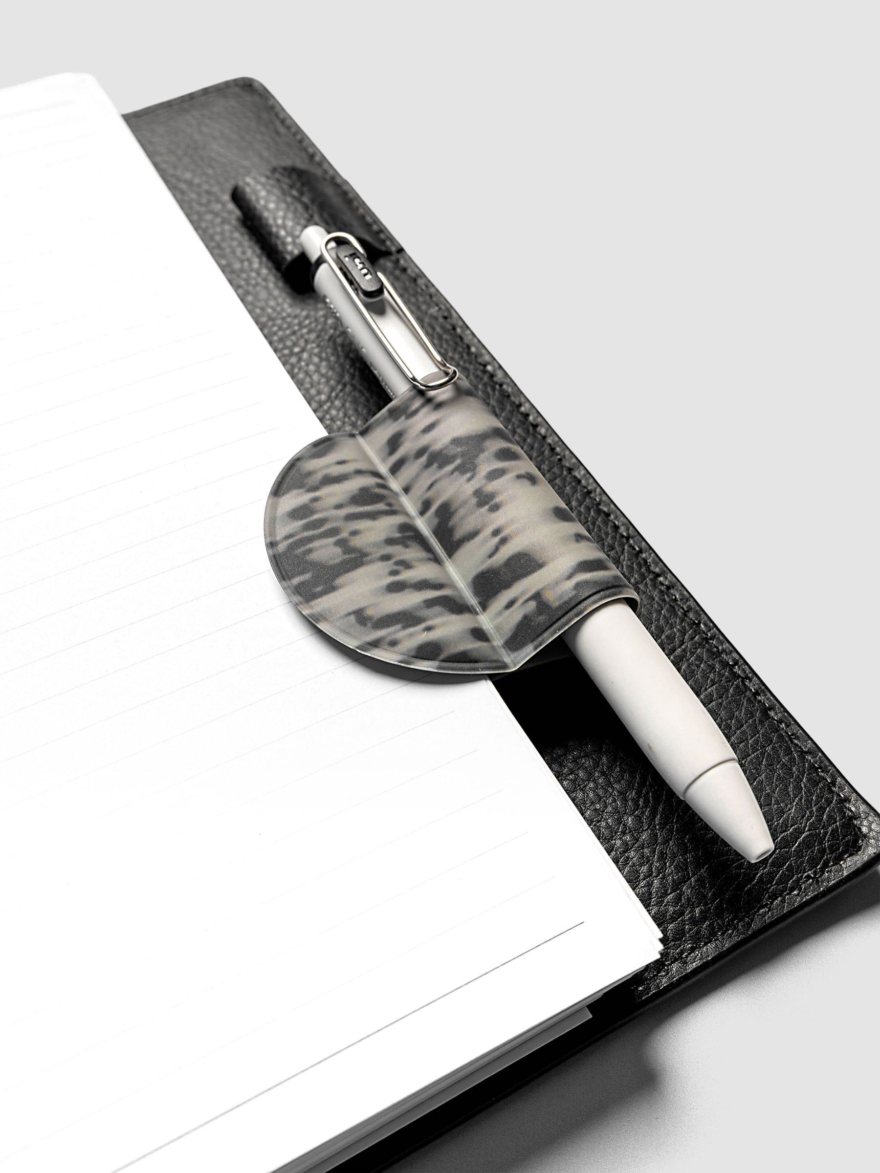 Magnetic Bookmark | Pen Holder | Vinyl in Blurred Ink holding a white pen, clipped to the spine of an open black leather planner