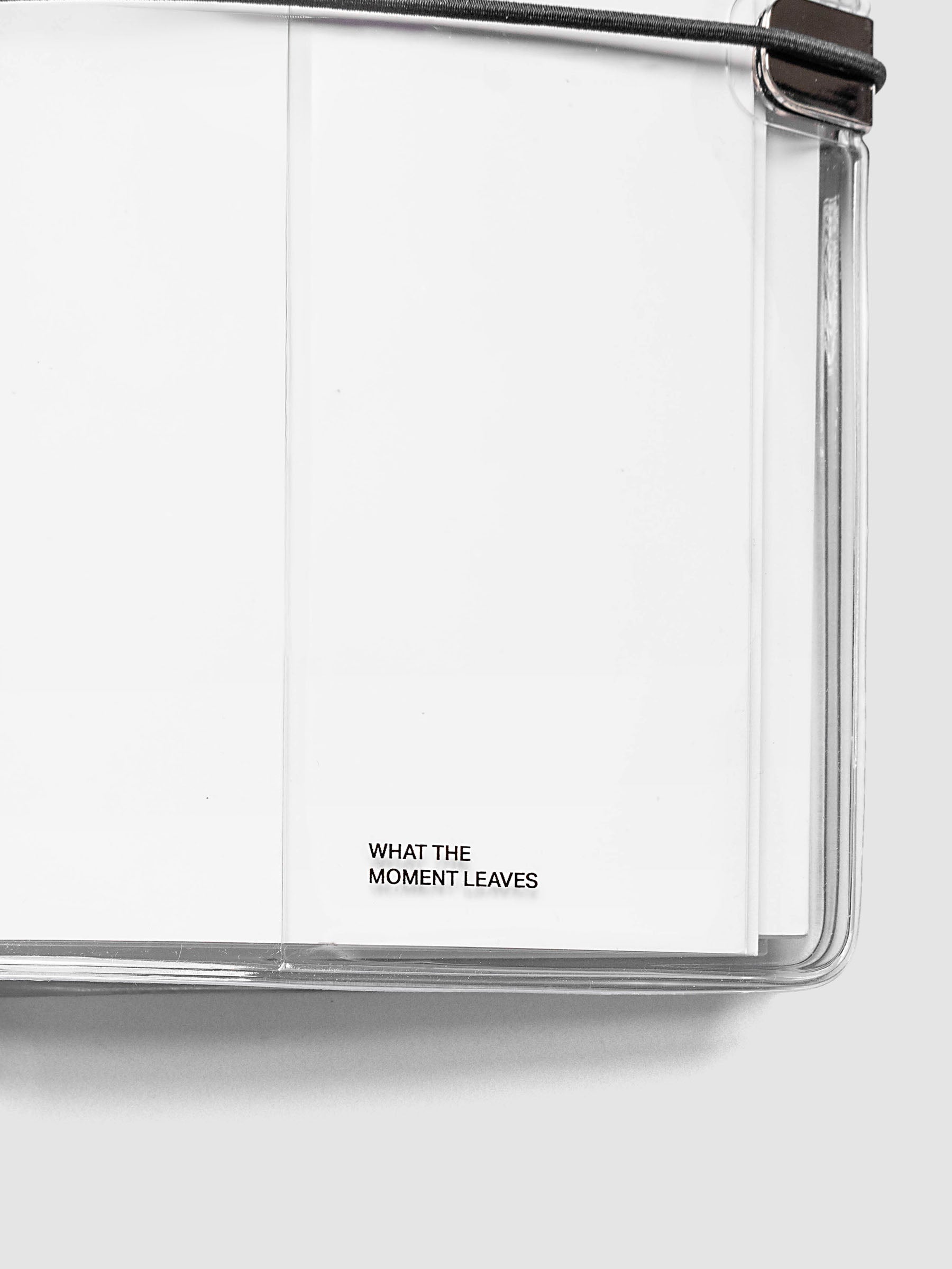 What Lasts Travel Notebook Cover in clear vinyl, A5 Slim, close-up of the bottom right corner with a white notebook loaded inside, showing the silver metal closure and black bungee cord detail