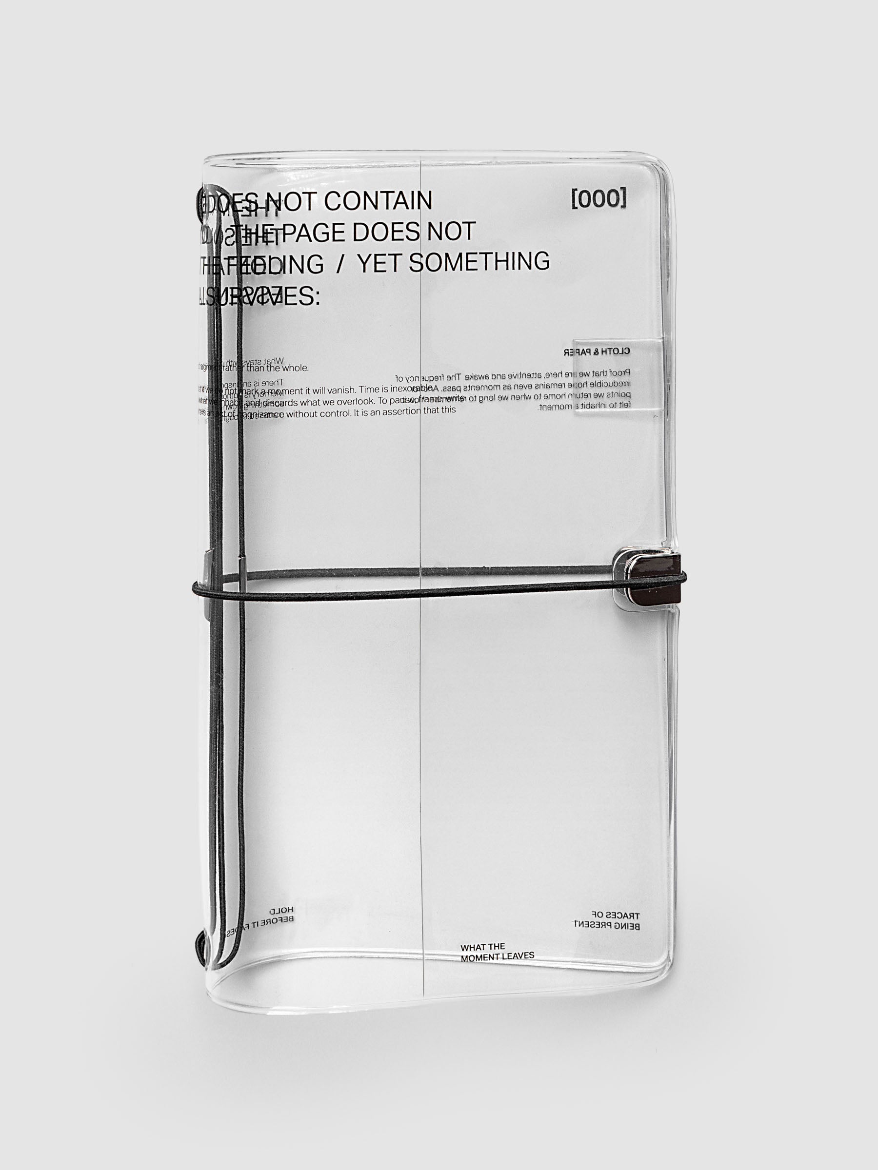 What Lasts Travel Notebook Cover in clear vinyl, A5 Slim, front exterior view, closed and upright against a light gray background, showing black typographic text printed on the vinyl and a black bungee cord with silver metal closure