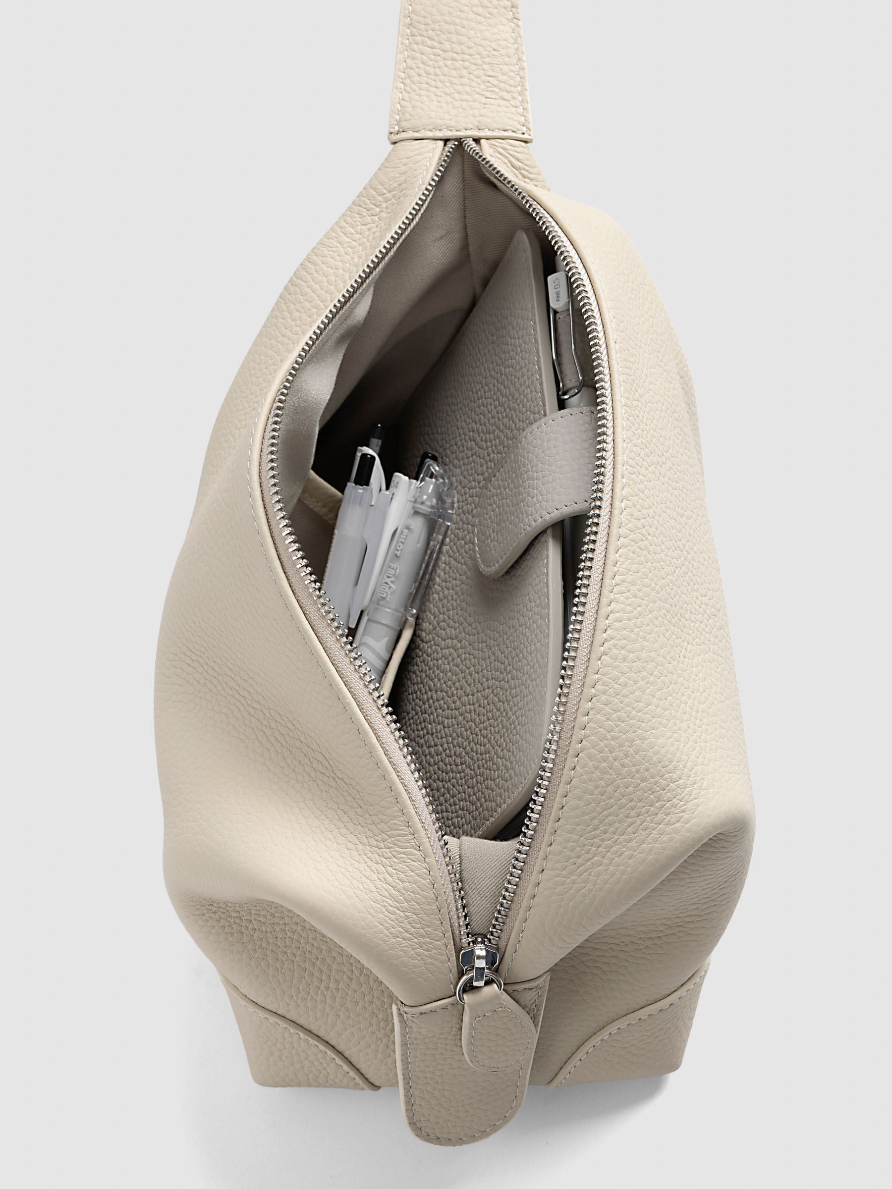 Folded Form Pouch in warm beige pebbled leather, top-down angled view, open and filled with pens and a notebook in the interior side pocket