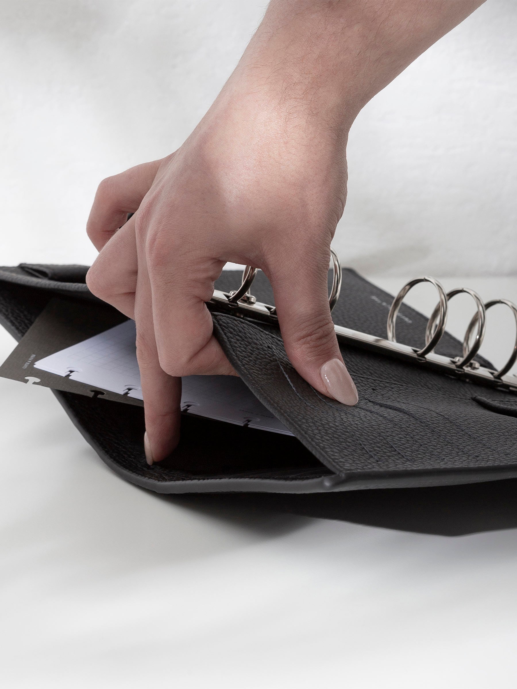A fair-skinned hand shown opening the back pocket of the black leather agenda on an off-white background. There are inserts and a dashboard inside the pocket.