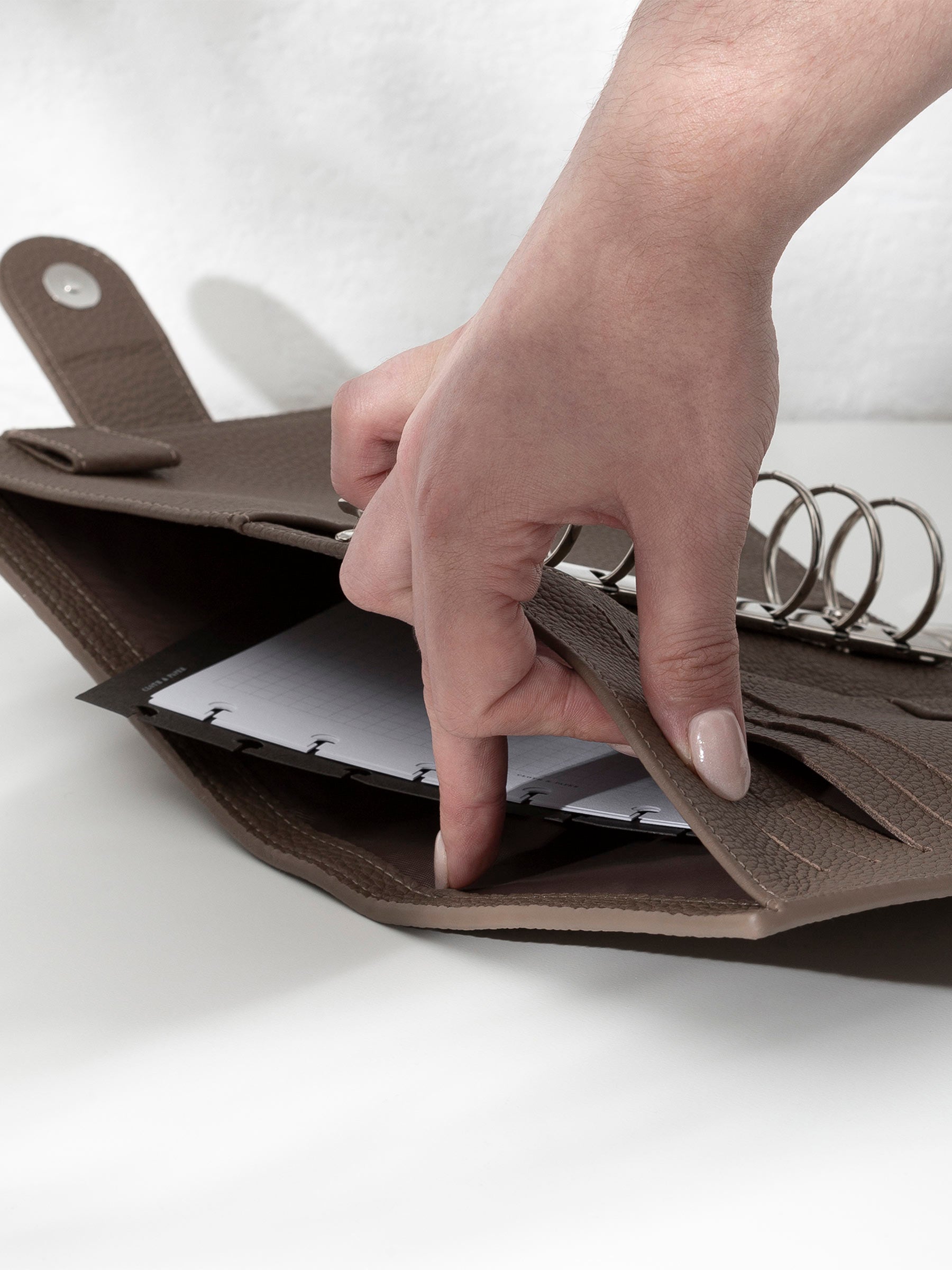 A fair-skinned hand shown opening the back pocket of the cortado agenda on a white background. There are inserts and a dashboard inside the pocket.