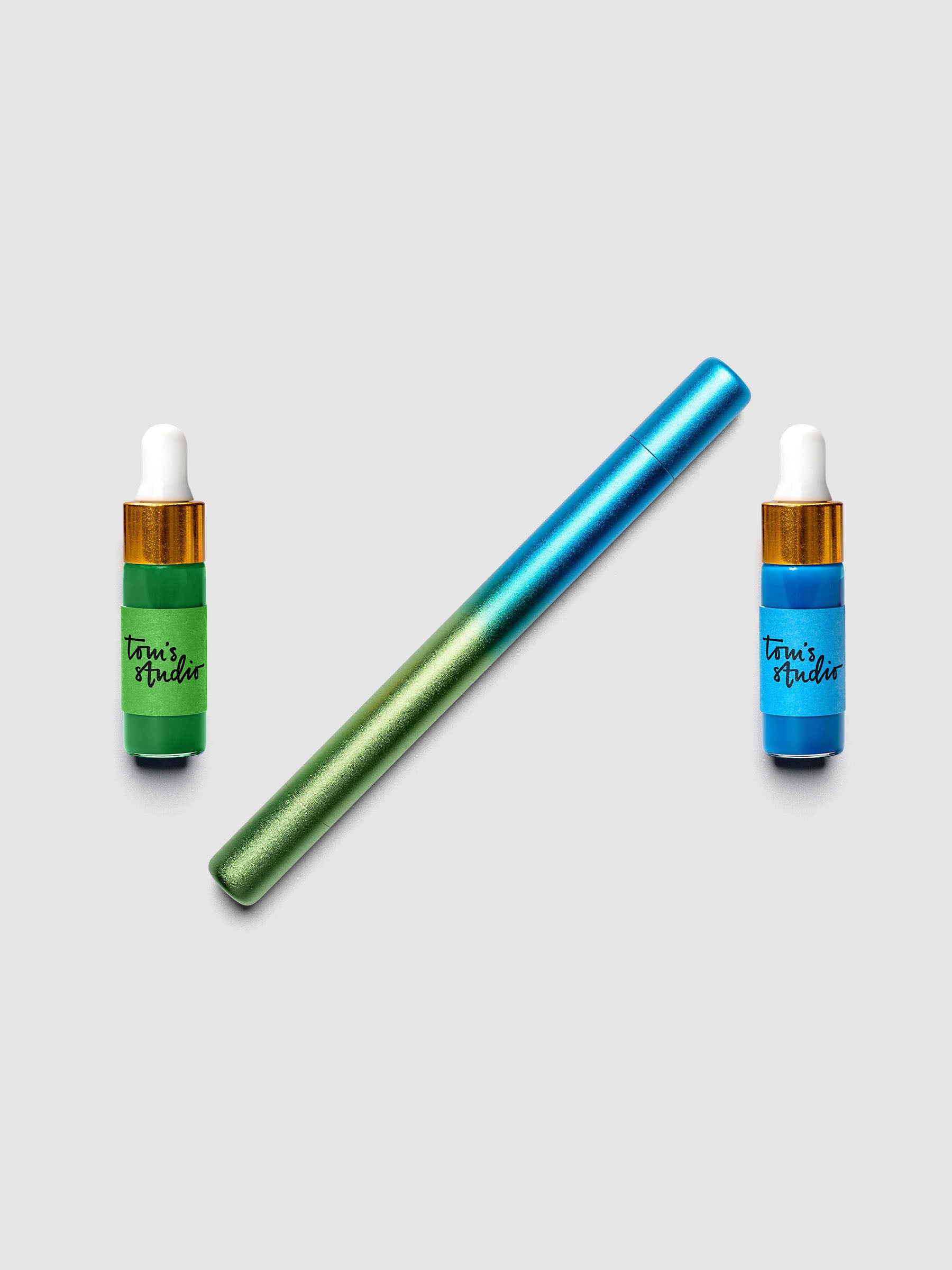 Tom's Studio Ori Refillable Highlighter in blue and green ombre finish laid diagonally between two ink bottles in matching blue and green