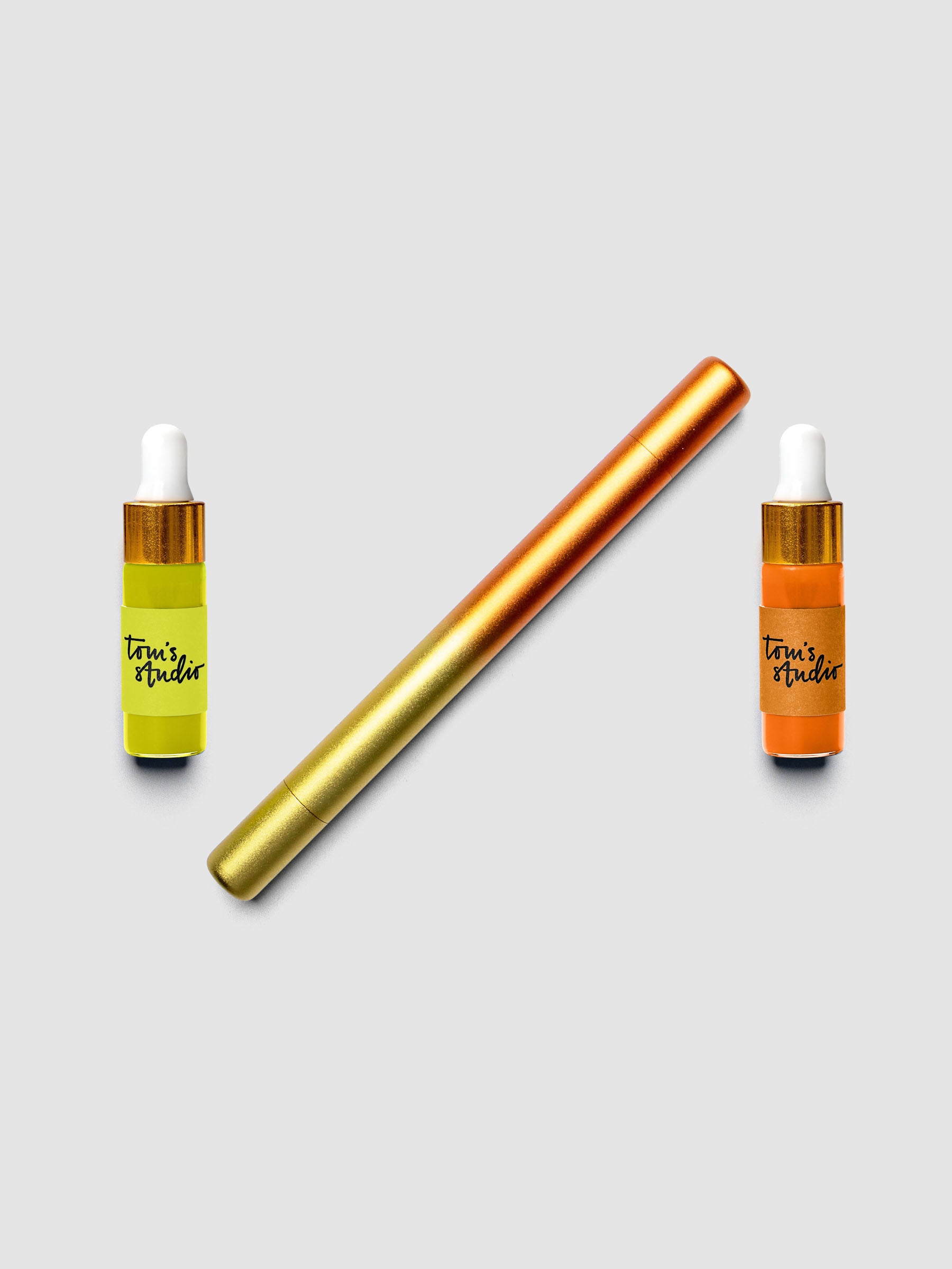Tom's Studio Ori Refillable Highlighter in orange and gold ombre finish laid diagonally between two ink bottles in matching yellow and orange