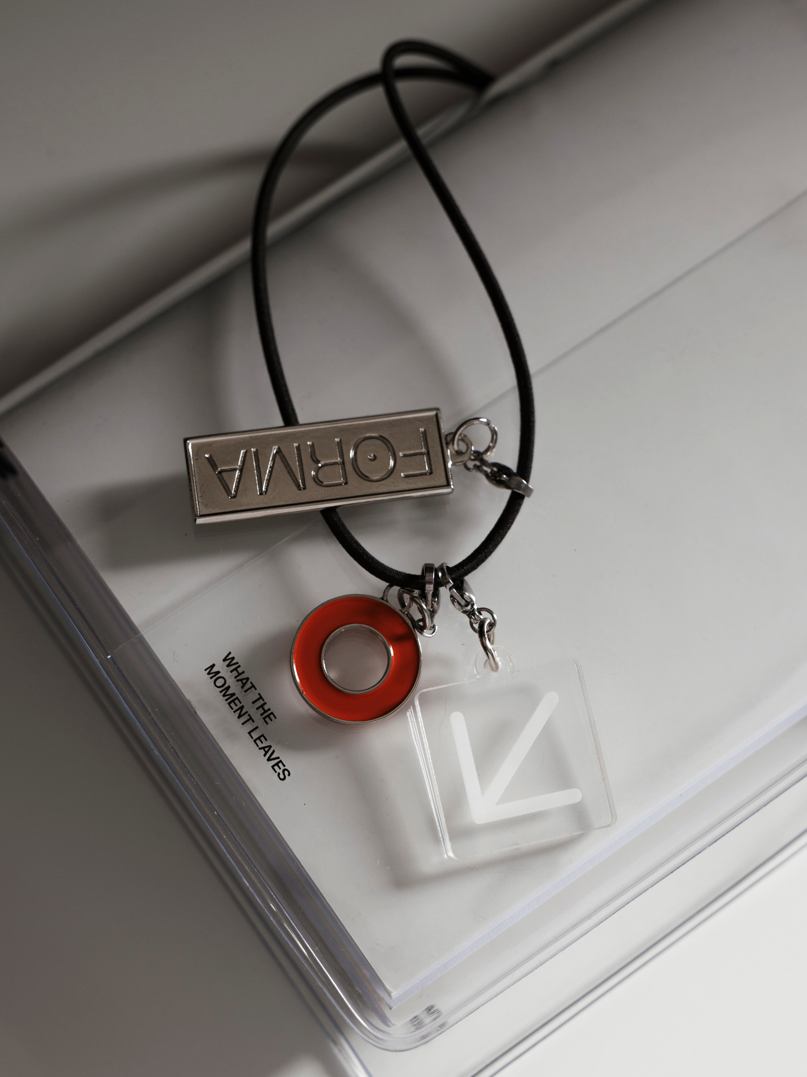 Travel Notebook Charms close-up on travel notebook exterior band showing SPOTLIGHT in deep orange enamel, FORMA in silver metal, and ARROW in translucent white plastic