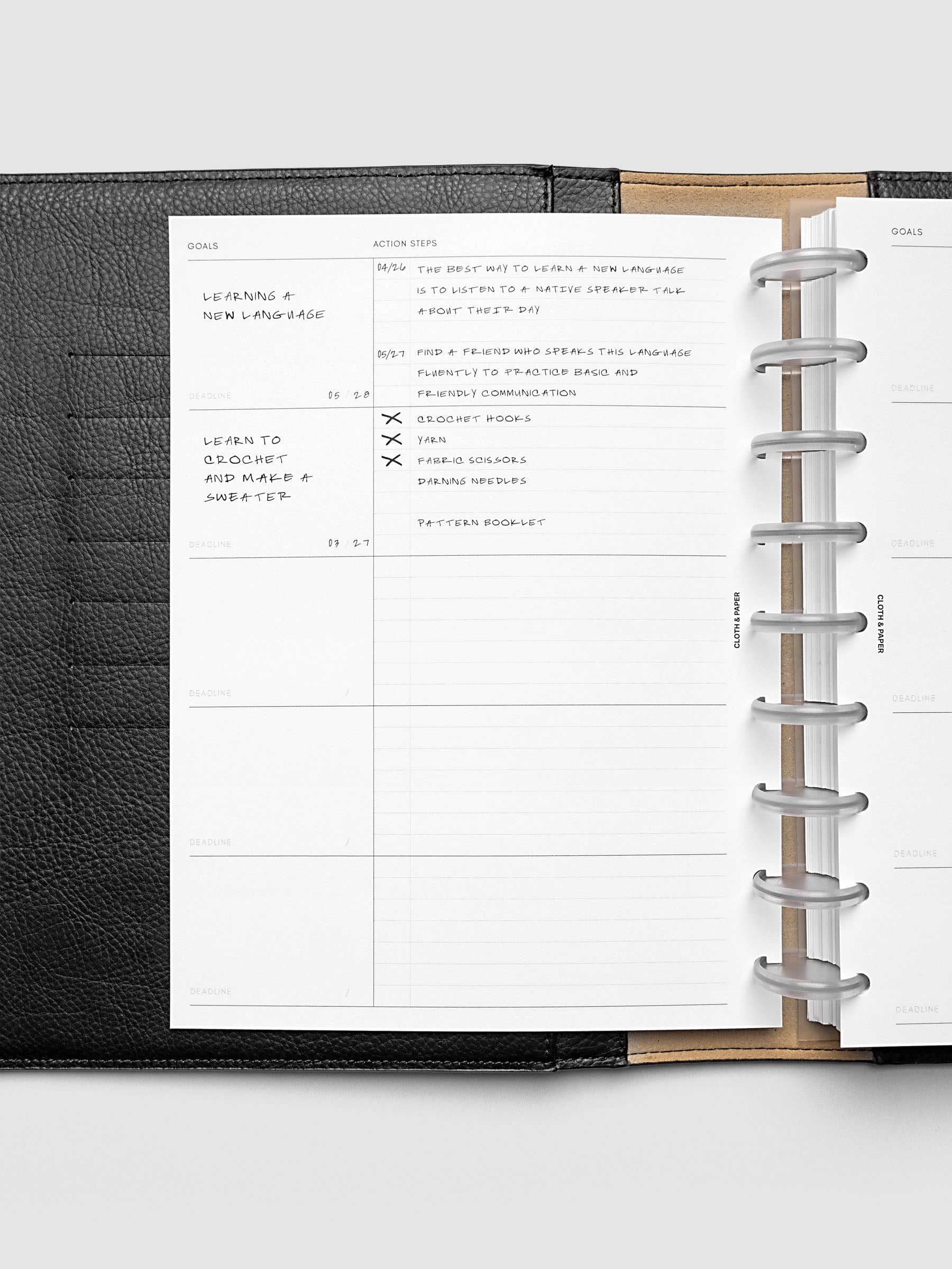 Open planner showing a goals page with handwritten notes about learning a new language and knitting a sweater, alongside a list of action steps. The planner is bound with metal rings and has a black leather cover.