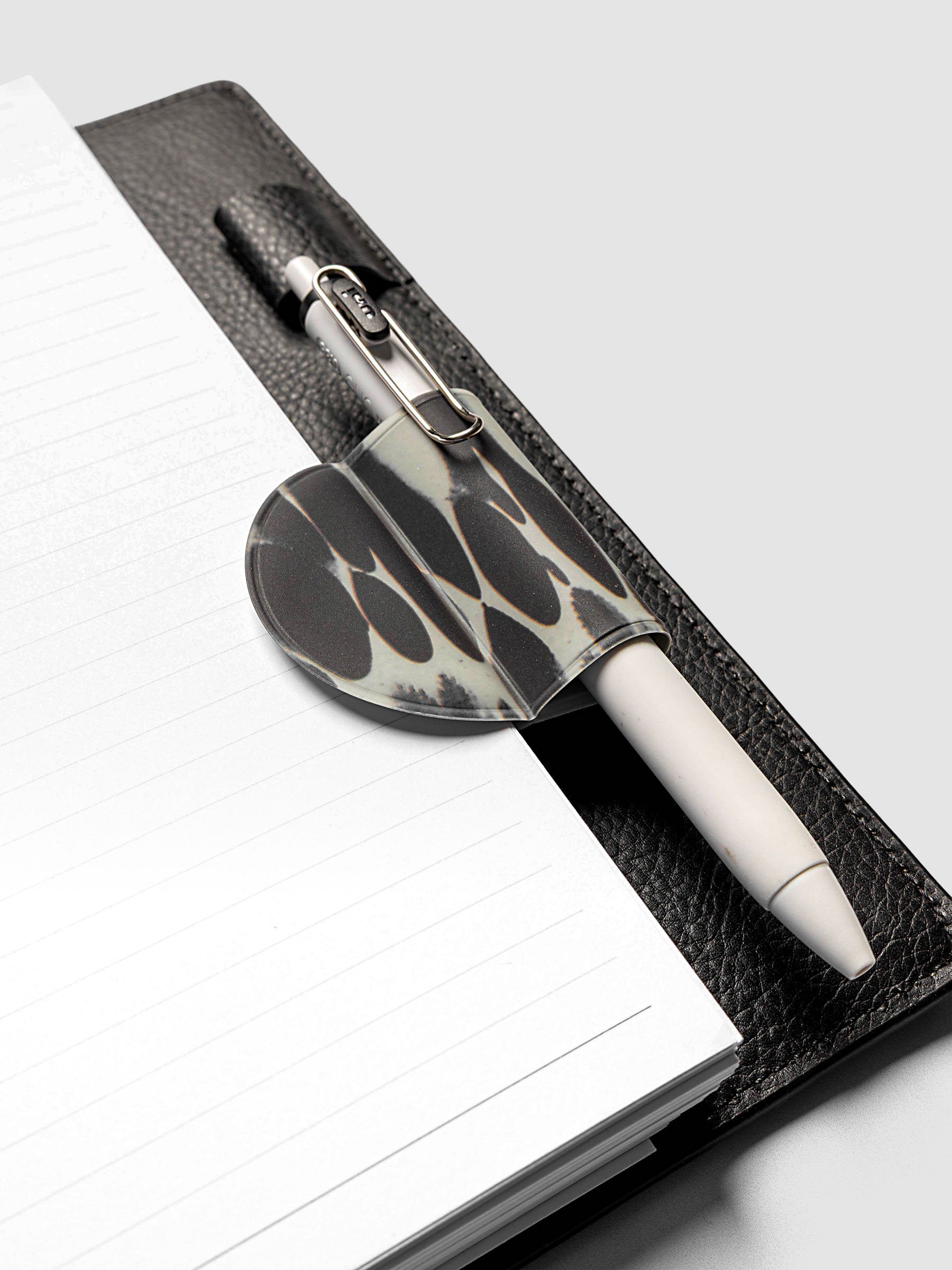 Magnetic Bookmark | Pen Holder | Vinyl in Dissolve holding a white pen, clipped to the spine of an open black leather planner
