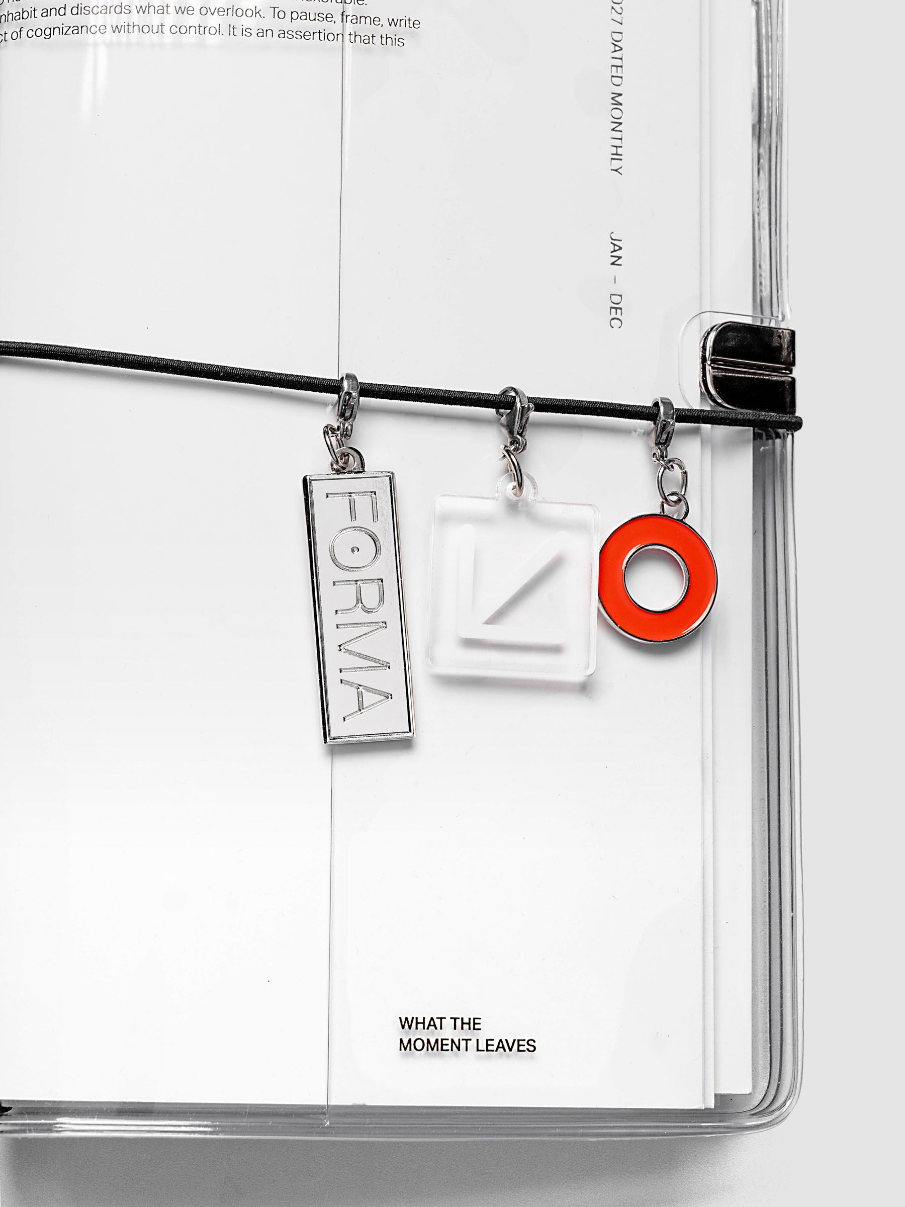 Travel Notebook Charms on exterior band of a closed travel notebook showing SPOTLIGHT, FORMA, and ARROW charms attached via lobster clasp