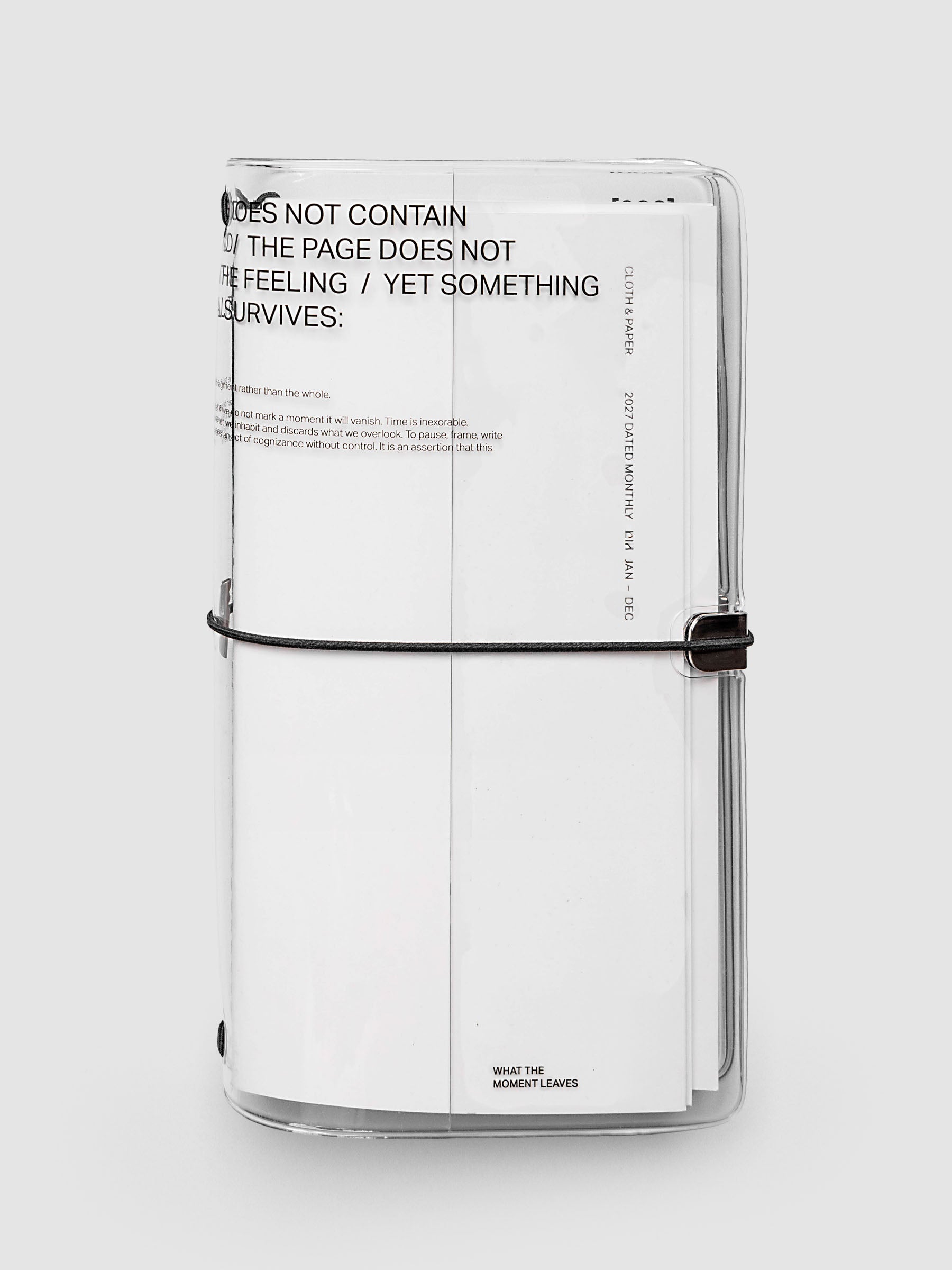 What Lasts Travel Notebook Cover in clear vinyl, A5 Slim, front exterior view, closed and upright with a white notebook loaded inside, showing typographic text on the vinyl and black bungee cord with silver metal closure