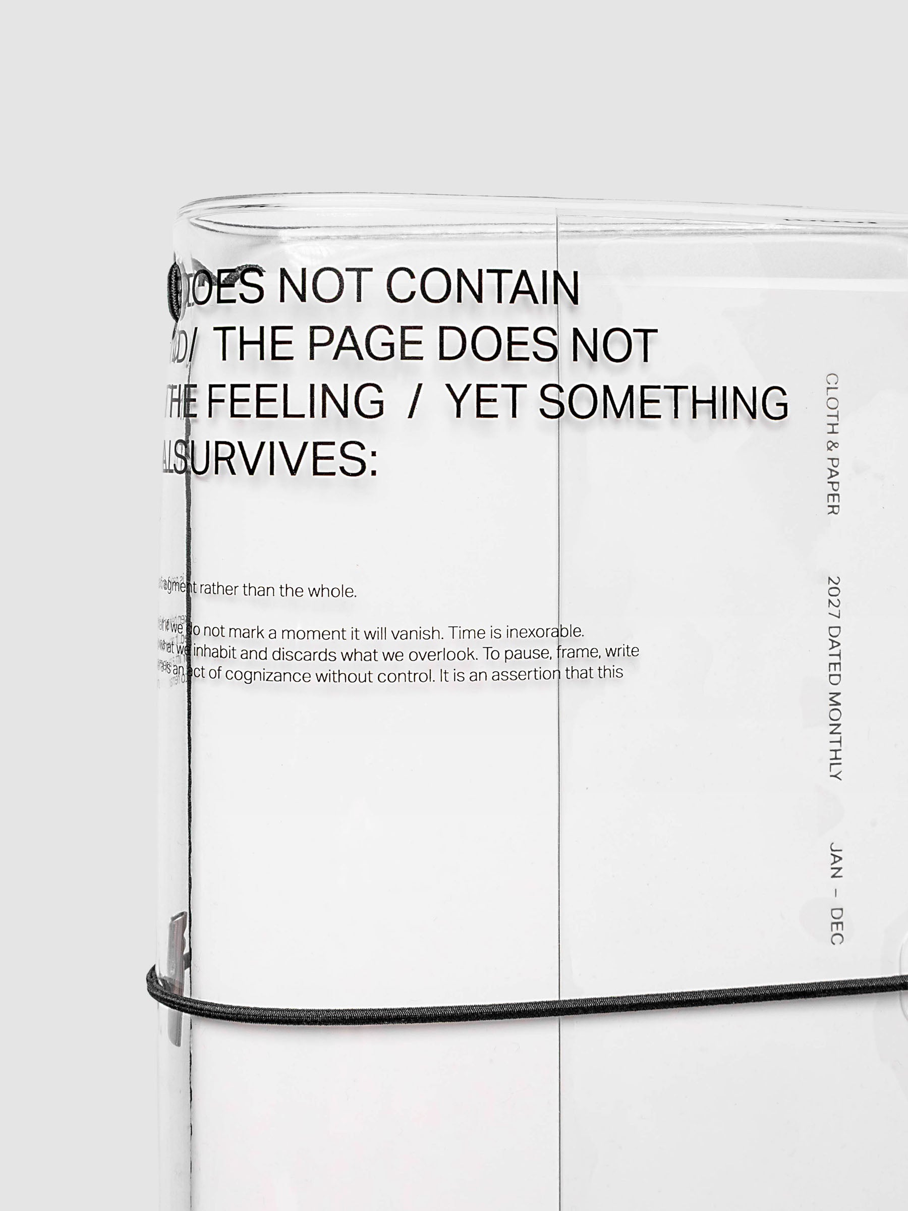 What Lasts Travel Notebook Cover in clear vinyl, A5 Slim, close-up of the upper portion showing black typographic text printed on the vinyl surface with a white notebook loaded inside and black bungee cord visible along the bottom edge