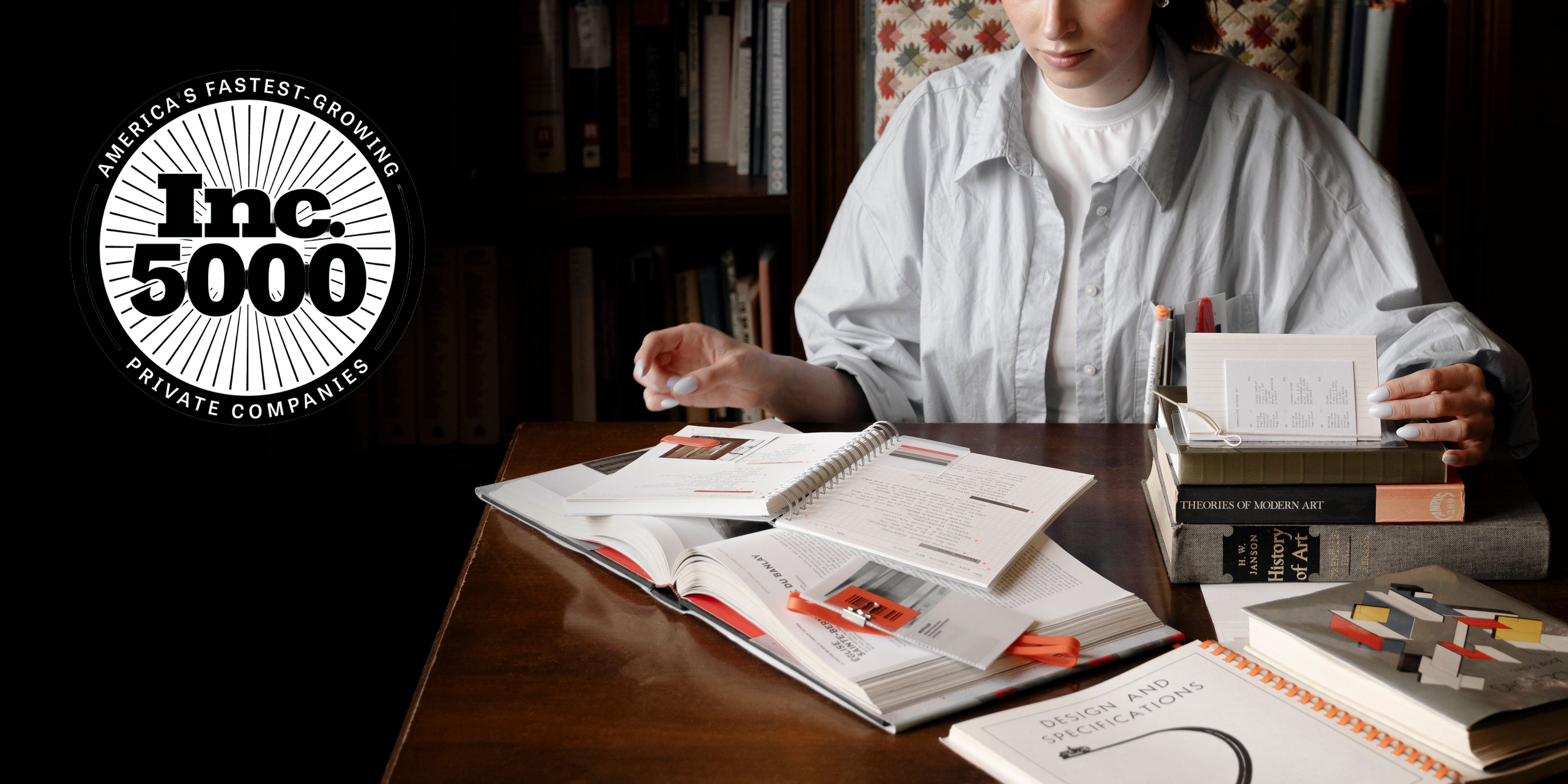 A woman is shown browsing through a pile of several books and notebooks displayed on a desk.  In the upper left corner is a logo for Inc. 5000 as Cloth & Paper was recognized for being one of America's Fastest-Growing Private Companies.