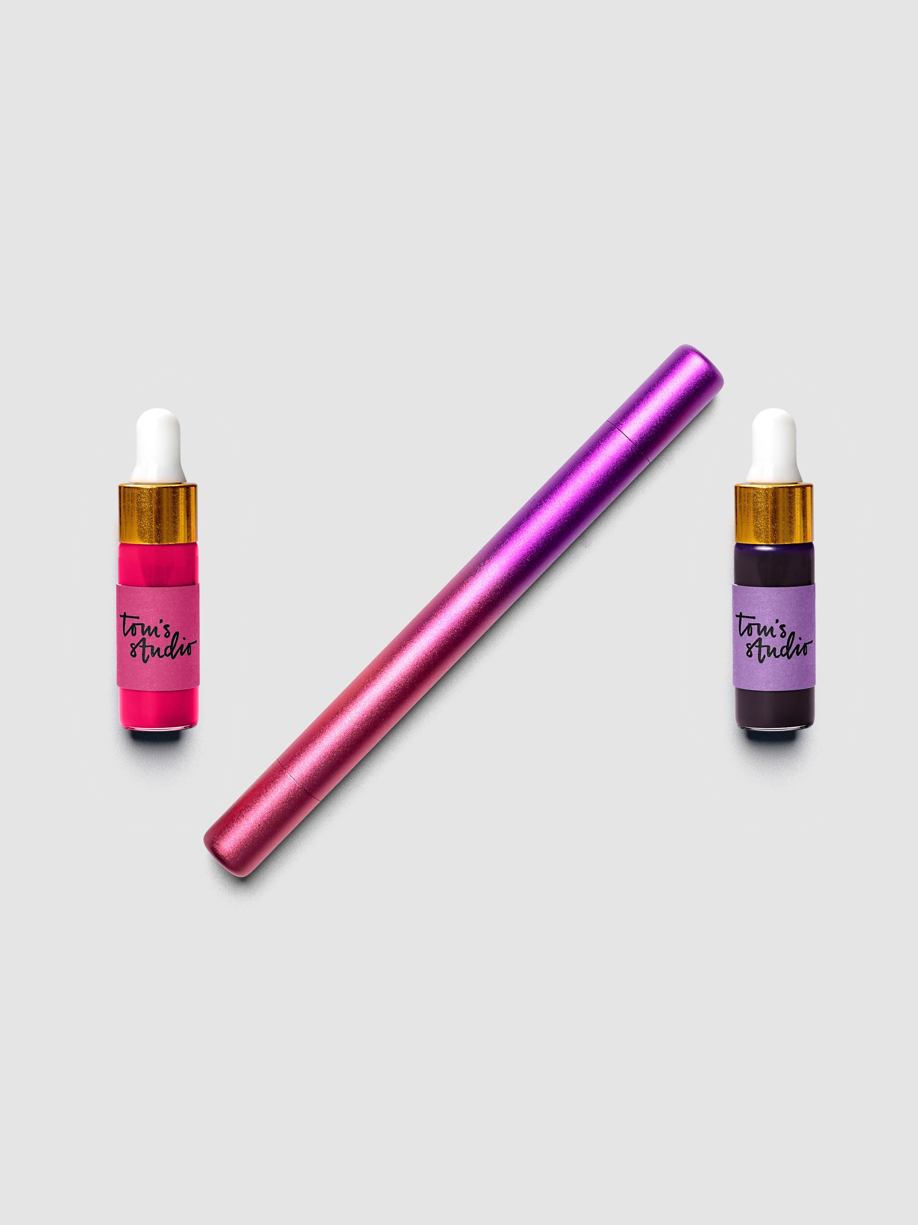 Tom's Studio Ori Refillable Highlighter in purple and pink ombre finish laid diagonally between two ink bottles in matching pink and purple
