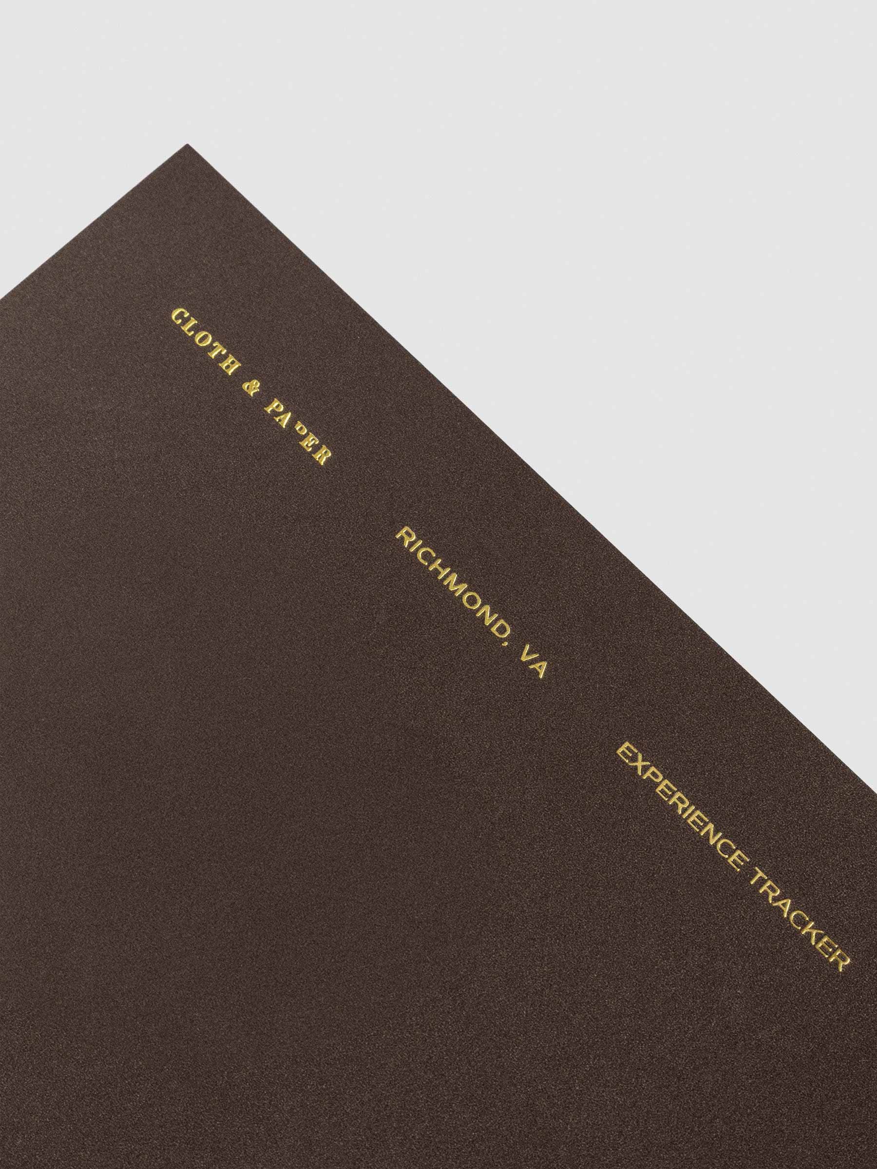 Close up image of the Experience Tracker Walnut Pocket Plus notebook from the Traveler's Notebook Collection. The cover has gold foil text that reads Cloth and Paper Richmond, VA Monthly Admin and is displayed on a neutral background